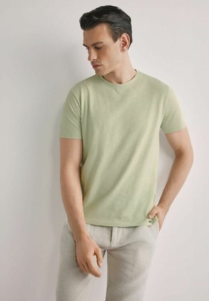 Mężczyzna w jasnozielonej koszulce z krótkim rękawem i beżowych spodniach, stojący z jedną ręką w kieszeni, patrzący w dół i na bok.