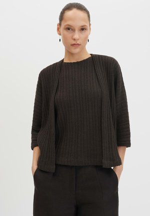 Frau trägt schwarzen gerippten Strickpullover und passenden Cardigan mit Dreiviertelärmeln, Hände in den Taschen der schwarzen Hose, neutraler Hintergrund.
