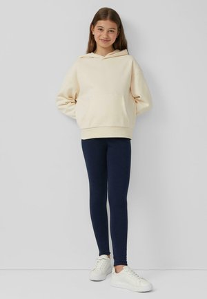 Felpa crema con tasca frontale, abbinata a leggings blu navy e sneakers bianche. Texture liscia, design casual e vestibilità rilassata.