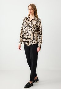 Crème en zwarte blouse met zebraprint, een gekraagde halslijn en lange mouwen, gecombineerd met zwarte pantalon en loafers.