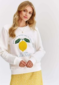 Bílá mikina s texturovaným žlutým citronovým grafickým prvkem a modrým textem na přední straně. Spárována se sukní s žlutými puntíky.