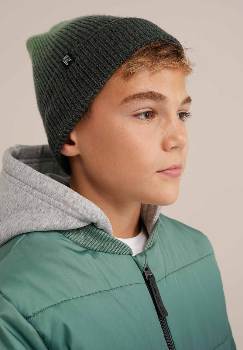 Green ribbed knit beanie on a person, paired with a gray hoodie and teal puffer jacket. Features a black logo tag on the beanie.