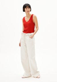 Rode geribde tanktop en losse witte broek met trekkoord, met zijzakken. Model staat in zachte verlichting tegen een witte achtergrond.