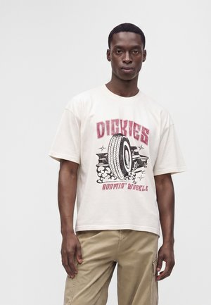 Άνδρας που φοράει λευκό T-shirt Dickies με γραφικό ελαστικού και μπεζ cargo παντελόνι, στέκεται μπροστά σε απλό φωτεινό φόντο.
