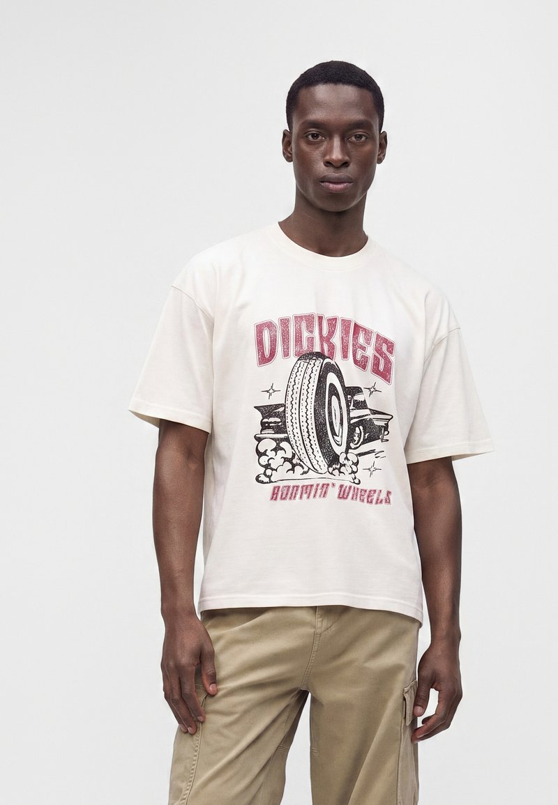 Άνδρας που φοράει λευκό T-shirt Dickies με γραφικό ελαστικού και μπεζ cargo παντελόνι, στέκεται μπροστά σε απλό φωτεινό φόντο.