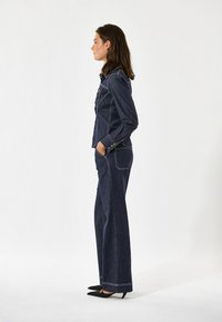 Jeansowa kurtka z dopasowanymi spodniami w szerokie nogawki w ciemnoniebieskim kolorze, z białymi przeszyciami, guzikami i bocznymi kieszeniami.