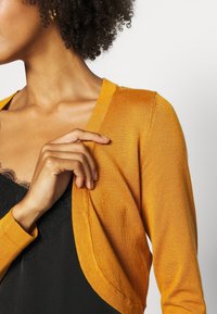 Senfgelbes, leichtes, gestricktes Cardigan mit V-Ausschnitt und offener Vorderseite, getragen über einem schwarzen Oberteil mit Spitzenbesatz, das eine glatte Textur zeigt.