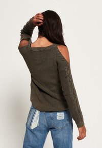 Olivově zelený pletený svetr s výřezy na ramenou, textura rukávů s kabelkovým pletením a volný střih. Nosený s modrými roztrhanými džíny.