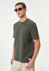 Oliven grøn textureret T-shirt med korte ærmer og rund halsudskæring, med et lille hvidt mærke i nederste venstre hjørne.
