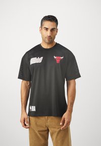 Crna košarkaška majica s kratkim rukavima od mrežaste tkanine s crvenim logotipom bika i bijelim tekstom "NBA". Dizajn uključuje teksturiranu tkaninu i širok kroj.