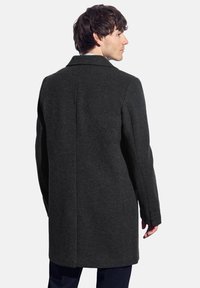 Muž stojící bokem, oblečený v tmavě šedém vlněném kabátu ke kolenům přes tmavé kalhoty, proti jednolitému světlému pozadí.