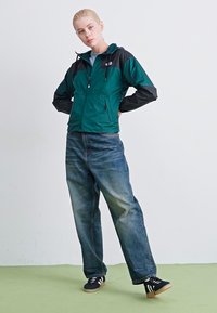 Groene en zwarte zip-up jack met een capuchon; gemaakt van ademend, waterafstotend materiaal. Gekocht in combinatie met losse blauwe jeans en zwarte sneakers.