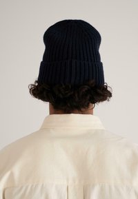 Dark blue knitted beanie with ribbed texture, featuring a folded brim. Worn by a person with curly hair, view focuses on the back of the beanie.