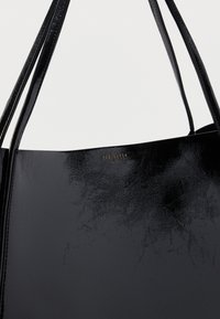 Fekete műbőr táska fényes felülettel, dupla fogantyúval és aranyozott, dombornyomott logóval a felső középső részen.