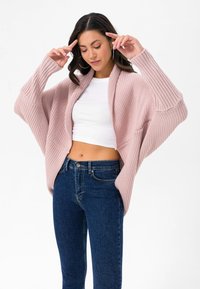 Hellrosa, übergroßer Cardigan mit gerippter Textur, offener Vorderseite und tiefen Taschen, kombiniert mit eng anliegenden blauen Jeans.