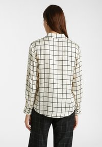 Camicia a maniche lunghe in tessuto crema con un motivo a griglia nera, dotata di colletto e polsini abbottonati. L'orlo è leggermente arrotondato sul retro.