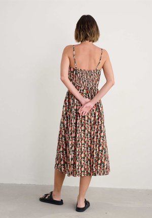 Frau steht barfuß auf einem schlichten Boden und trägt ein ärmelloses, florales Midikleid mit gerafftem Rücken, die Hände hinter dem Rücken verschränkt.