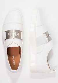 Vita slip-on sneakers med en slät syntetisk ovandel, elastisk rem och metalllogotyp som accent. Har en vadderad innersula och vit gummisula.