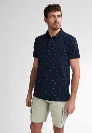 Marineblaues Poloshirt mit kleinem, mehrfarbigem Fischmuster, kurzen Ärmeln und einem Drei-Knopf-Verschluss, kombiniert mit hellgrünen Cargo-Shorts.