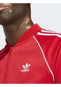 Červená sportovní bunda na zip s bílými pruhy, rebrovaným límcem a vyšívaným bílým logem Adidas na hrudi. Hladká textura látky.