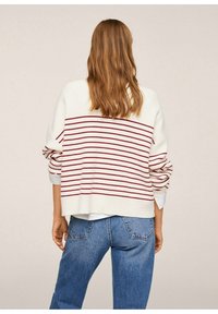 Persona con lunghi capelli castano chiaro che indossa un maglione bianco con strisce orizzontali rosse e jeans blu, girata di spalle su uno sfondo neutro.