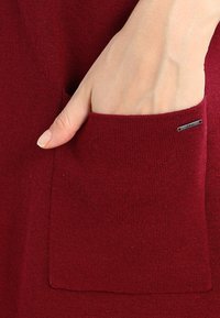Bordový pletený oděv s boční kapsou a elegantním kovovým akcentem. Materiál má jemnou texturu a hladký povrch.