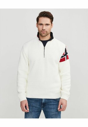 Egy férfi fehér, texturált, félcipzáras pulóvert visel, amelynek bal ujján norvég zászló mintázat látható, valamint kék farmert, és egyszerű háttér előtt áll.