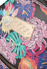 Sac à dos coloré à motif floral en matériau synthétique, avec une plaque logo dorée, une base noire et un compartiment zippé.