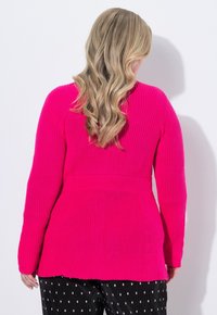 Blond włos z tyłu, ubrany w jasnoróżowy sweter w prążki i czarne spodnie w białe kropki, na białym tle.