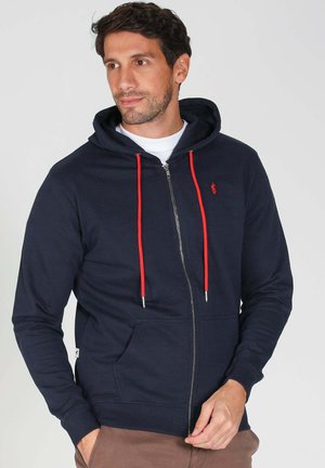 Sweat à capuche zippé de couleur marine en coton, avec des cordons de tirage rouges, des poches avant et un petit logo sur la poitrine. Texture lisse, design décontracté.