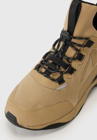 Tan athletic shoe featuring a textured upper, black laces, reflective accents, and a cushioned sole with a slightly raised heel.