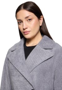 Manteau en mélange de laine gris avec une finition texturée, de grands revers et une coupe décontractée, porté sur un haut noir. Aucune fermeture visible.