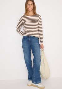 Gestreiftes Langarmshirt in Braun und Weiß, kombiniert mit blauen Schlagjeans. Das Model hält eine hellbeige Tasche. Sneakers mit Akzenten.