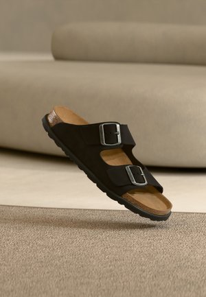 Zwarte sandaal met twee verstelbare gespen en kurk voetbed zwevend boven een beige tapijt voor een crèmekleurige bank.