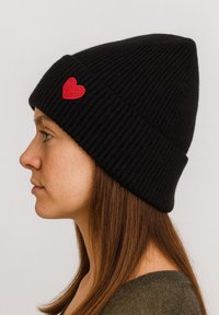 Woman in profile wearing a black ribbed knit beanie with a red embroidered heart on the cuff and a green top.
