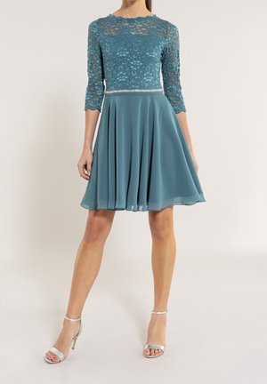 Kvinde iført teal knælængde kjole med blonder i toppen og flydende nederdel, kombineret med sølvfarvede højhælede sandaler.