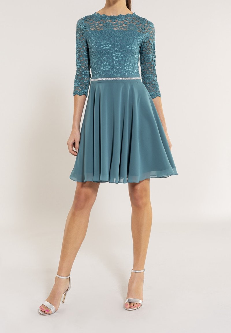 Femme portant une robe bleu sarcelle mi-longue avec un haut en dentelle et une jupe fluide, associée à des sandales argentées à talons hauts.