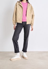 Beżowa kurtka z kapturem, różowy sweter, ciemne dżinsy i białe trampki z beżowymi akcentami. Posiada elastyczne materiałowe mankiety i luźny krój.