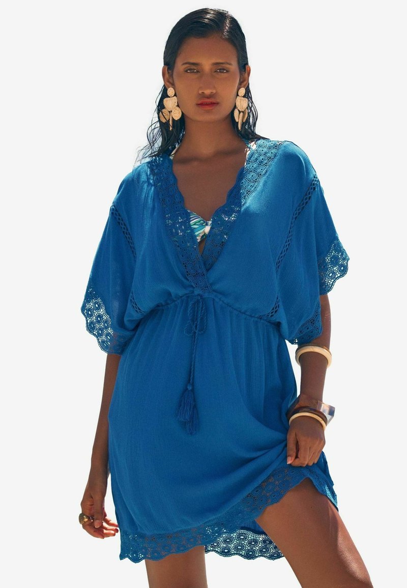 Blauwe kaftan van lichtgewicht stof, met kantafwerking, een V-halslijn met een strik en korte vleermuismouwen. Accenten zijn onder andere franjes.