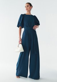 Teal wijde jumpsuit met pofmouwen, een getailleerde taille en een bijpassende strik. Gecombineerd met een kleine witte handtas met een gouden handvat.