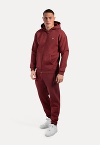 Felpa a zip color bordeaux con tasche frontali e pantaloni abbinati. Sneakers bianche con suole testurizzate. Tessuto morbido, vestibilità rilassata, branding minimale.