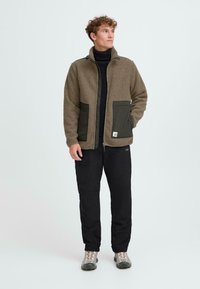 Giacca inpile verde oliva con finitura testurizzata, dotata di tasche verde scuro, chiusura con zip e vestibilità rilassata. Indossata con pantaloni cargo neri.