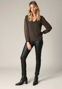 Langsleeve, V-hals top in metallic bruin met een geweven patroon, gecombineerd met strakke zwarte leren broek en enkellaarzen, staande pose.