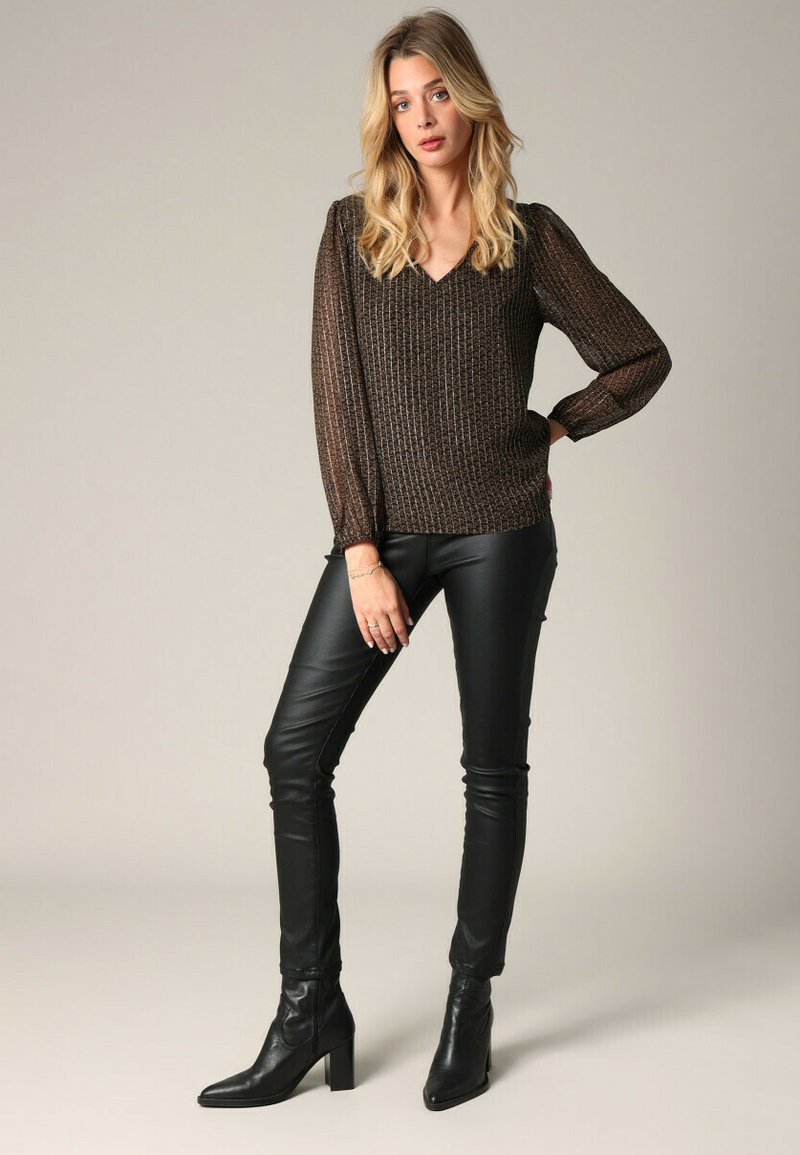 Langsleeve, V-hals top in metallic bruin met een geweven patroon, gecombineerd met strakke zwarte leren broek en enkellaarzen, staande pose.