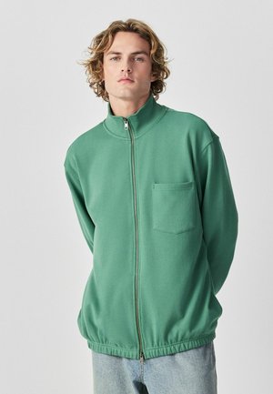 Junger Mann mit lockigem Haar, der eine grüne Jacke mit Reißverschluss und Brusttasche sowie hellblaue Jeans trägt und vor einem einfarbigen Hintergrund steht.