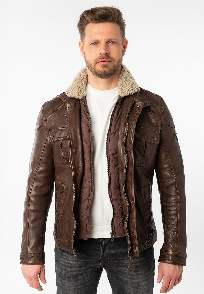 Braune Lederjacke mit Schafslederkragen, Reißverschluss, zwei Fronttaschen und einer gesteppten Innenseite. Über einem weißen Hemd getragen.