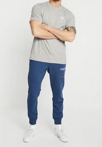 Grå bomullströja med en liten vit logotyp på bröstet, kombinerad med marinblåa sweatpants med en röd accentficka och vita sneakers.