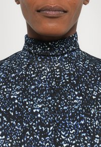 Top negru cu guler înalt, dintr-un material texturat, cu un model abstract în nuanțe de albastru și alb, cu detalii zimțate de-a lungul cusăturilor.