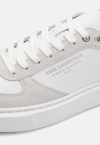 Baskets en suède gris clair et cuir blanc avec des lacets blancs, présentant une semelle texturée et un détail de logo en relief sur le côté.