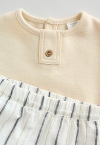 Beżowa bluza z guzikiem i prążkowanym dekoltem; na dole znajduje się materiał w białe paski z granatowymi i beżowymi pasami.